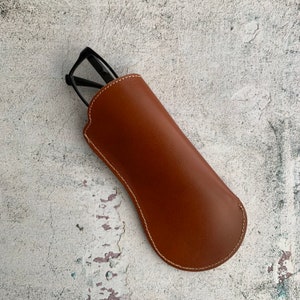Puede incluir: Un estuche de gafas de cuero marr&oacute;n con un borde cosido. El estuche est&aacute; vac&iacute;o y tiene una forma ligeramente curvada.