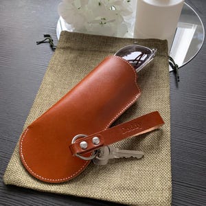 Puede incluir: Un estuche para gafas de cuero marr&oacute;n con un llavero y una correa que dice "Daddy".