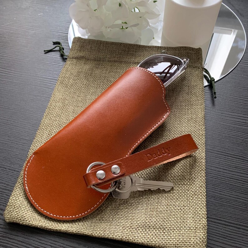 Puede incluir: Un estuche para gafas de cuero marr&oacute;n con un llavero y una correa que dice "Daddy".