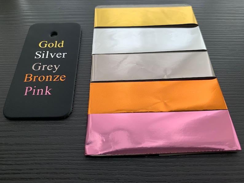 Puede incluir: Cinco l&aacute;minas met&aacute;licas de papel en diferentes colores: dorado, plateado, gris, bronce y rosa. Las l&aacute;minas est&aacute;n apiladas una encima de la otra, con la l&aacute;mina dorada en la parte superior y la l&aacute;mina rosa en la parte inferior.