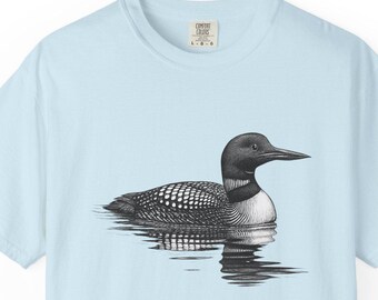 May include: Light blue t-shirt featuring a detailed black and white illustration of a loon floating on water. The bird has a dark head and back with a white-speckled pattern on its wings. The water's reflection is visible.