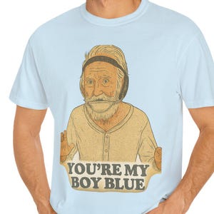 May include: Light blue t-shirt featuring a cartoon illustration of an elderly man with a mustache and headphones. The text "YOU'RE MY BOY BLUE" is printed below the illustration. The shirt is made of a soft material.