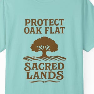 May include: A light blue t-shirt with the text "PROTECT OAK FLAT" above a brown tree graphic and the words "SACRED LANDS". The shirt is a short-sleeved crew neck.