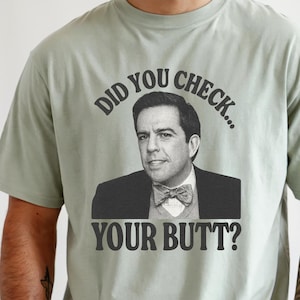 May include: A light green t-shirt featuring a black and white image of a man in a suit and bow tie. The text "DID YOU CHECK... YOUR BUTT?" is above and below the image. The shirt has a crew neck and short sleeves.