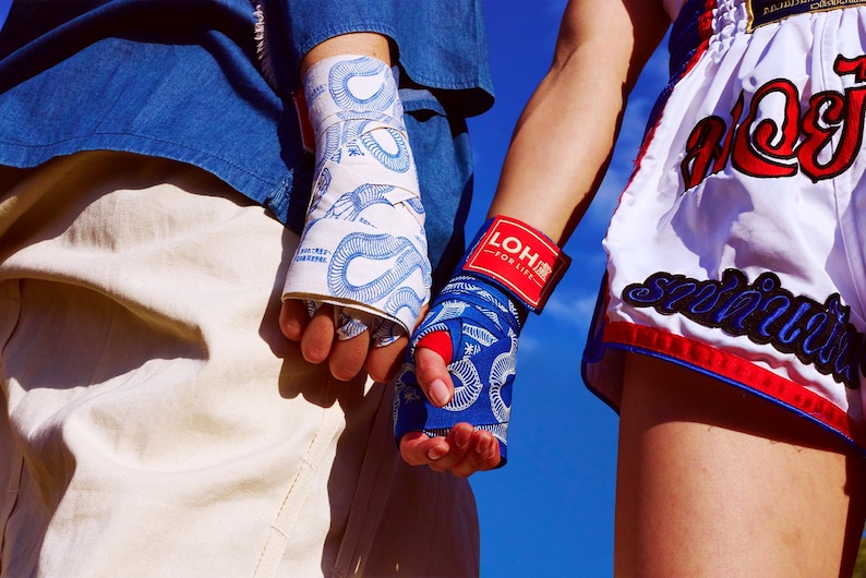 May include: Two pairs of boxing hand wraps, one white with blue snake pattern, the other blue with a red accent. The blue wrap has a red wristband with the text "LOH FOR LIFE". The image also shows white and red boxing shorts.