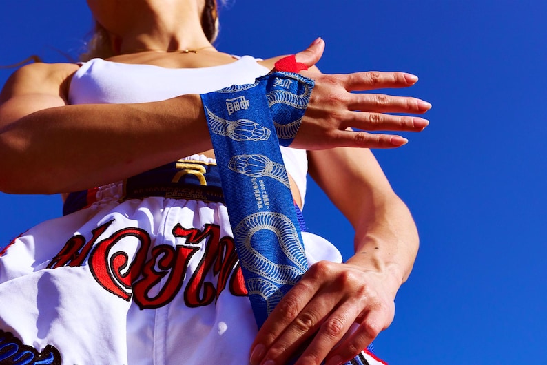 May include: A blue and white Muay Thai hand wrap with a snake design is wrapped around a wrist. The person is wearing a white top and a white Muay Thai shorts with red and gold lettering. The background is a clear blue sky.