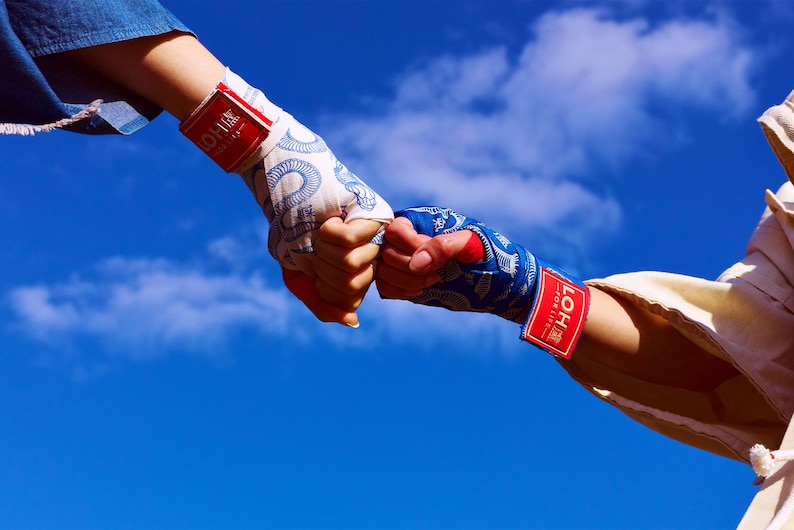 May include: Two fists bump, each wrapped in patterned hand wraps. One wrap is white with a blue design, the other is blue with a white design. Each wrap has a red label with white text. The background is a bright blue sky.