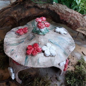 Puede incluir: Una casa de cerámica en forma de hongo con un techo blanco y verde, decorada con flores rojas y blancas. La casa está sentada sobre un lecho de musgo.