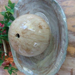 May include: A handcrafted ceramic bird bath featuring a light brown, spherical water basin and a wide, shallow, scalloped-edge dish in shades of green and brown. The bird bath is set on a wooden surface with artificial greenery.