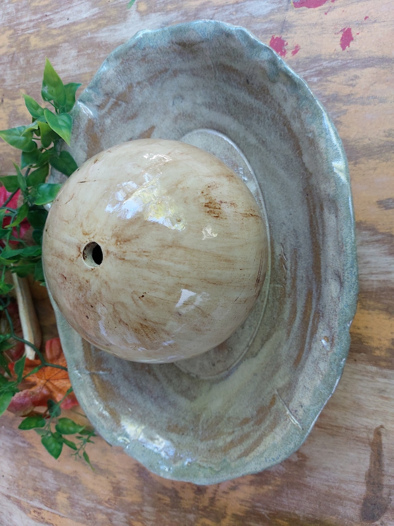 May include: A handcrafted ceramic bird bath featuring a light brown, spherical water basin and a wide, shallow, scalloped-edge dish in shades of green and brown. The bird bath is set on a wooden surface with artificial greenery.