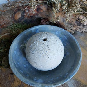 Könnte beinhalten: Eine weiße Keramikskugel mit gesprenkelter Textur und einem kleinen Loch oben, die in einer blauen und weißen Keramikschale mit gesprenkeltem Überzug ruht.