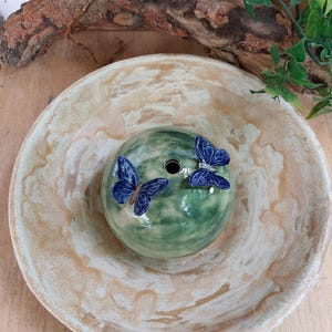May include: A ceramic decorative piece featuring a green, spherical form with two blue butterfly accents. The piece sits in a beige and white bowl. The design is suitable for home decor.