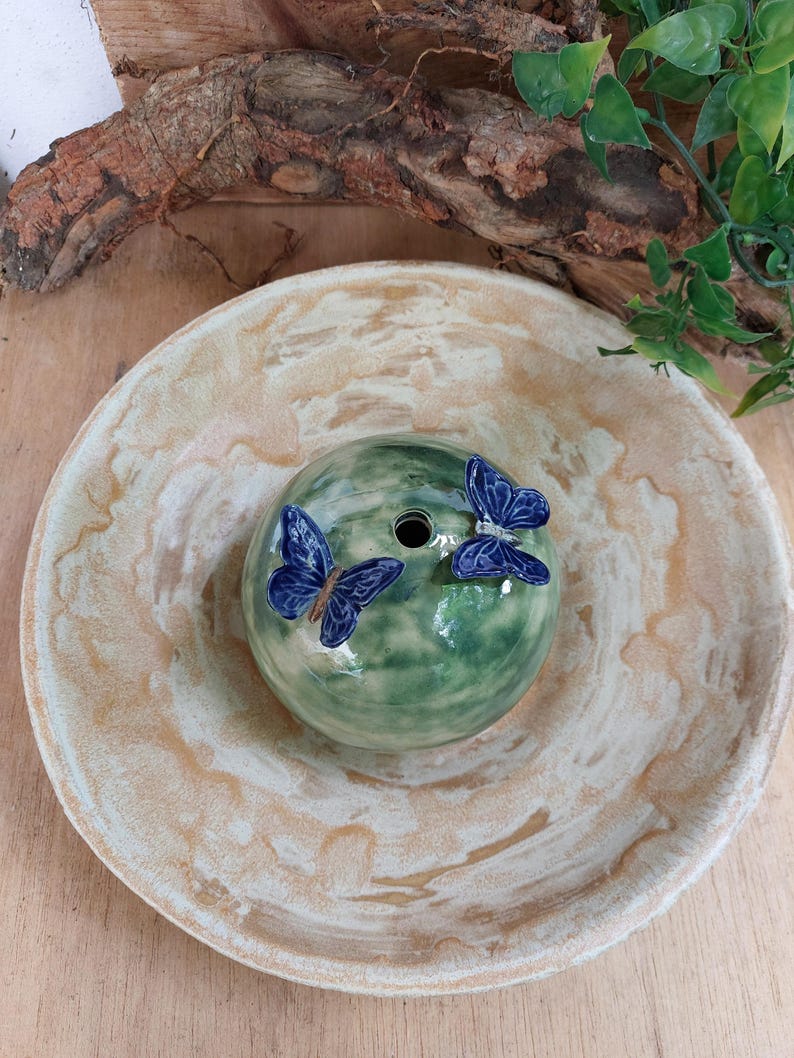 May include: A ceramic decorative piece featuring a green, spherical form with two blue butterfly accents. The piece sits in a beige and white bowl. The design is suitable for home decor.