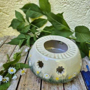 Könnte beinhalten: Ein cremefarbener Kerzenhalter aus Keramik, verziert mit Gänseblümchen-Ausschnitten und bemalten Gänseblümchen. Der Halter hat eine kreisförmige Öffnung oben und steht auf einer Holzoberfläche. Grüne Blätter und Gänseblümchen sind um den Halter angeordnet.