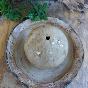 May include: A handmade ceramic bird bath with a round, textured bowl and a central, spherical water feature. The bird bath has a beige and brown colour scheme with decorative accents around the rim. The water feature has a hole in the centre.