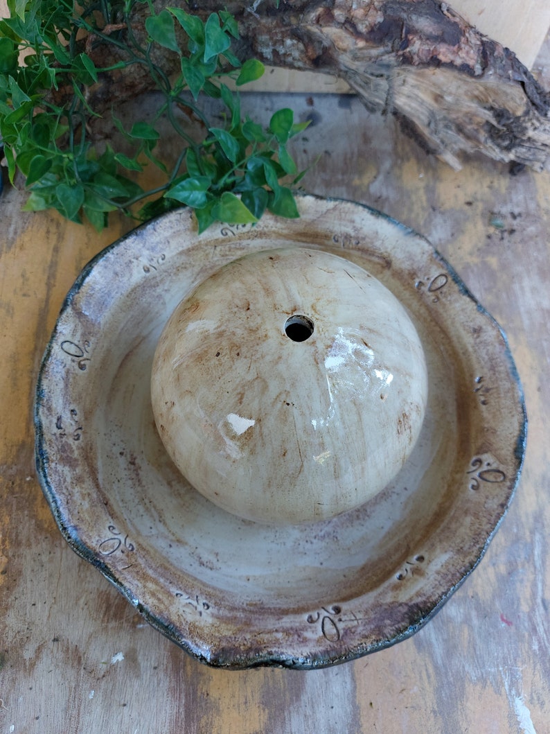 May include: A handmade ceramic bird bath with a round, textured bowl and a central, spherical water feature. The bird bath has a beige and brown colour scheme with decorative accents around the rim. The water feature has a hole in the centre.