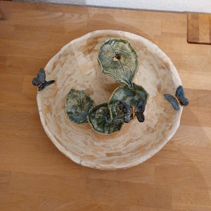 May include: A handmade ceramic bowl with a cream-coloured exterior and a textured surface. The bowl features three green leaf-shaped accents and three blue butterfly decorations. The bowl is a decorative piece.
