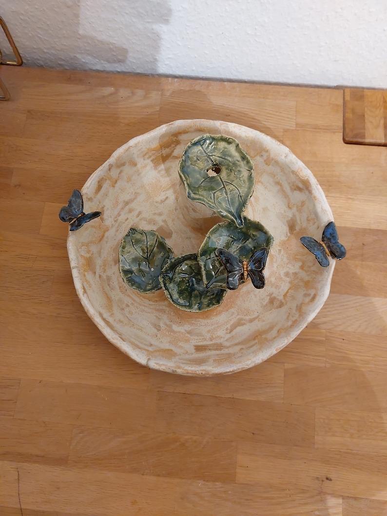 May include: A handmade ceramic bowl with a cream-coloured exterior and a textured surface. The bowl features three green leaf-shaped accents and three blue butterfly decorations. The bowl is a decorative piece.