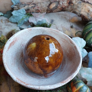 Könnte beinhalten: Eine glasierte Keramikkugel mit einem kleinen Loch oben, die in einer rustikalen, cremefarbenen Schale liegt. Die Kugel hat eine glänzende, braune Oberfläche mit dunkleren braunen Marmorierungen. Die Schale hat ein strukturiertes, gealtertes Aussehen.