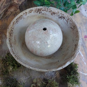 Könnte beinhalten: Ein handgefertigtes Keramik-Set aus Schale und Kugel. Die cremefarbene Schale hat ein Blumenmuster am Rand. Die Kugel ist gesprenkelt und hat ein kleines Loch. Das Set dient dekorativen Zwecken.