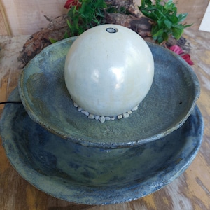 May include: A two-tiered ceramic water fountain. The base is a large, shallow bowl in shades of blue and green. A smaller bowl sits above it, supporting a spherical, off-white ceramic orb. Small stones surround the orb.