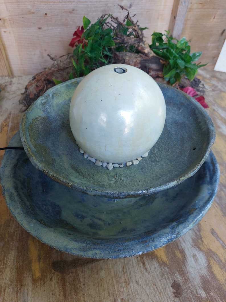 May include: A two-tiered ceramic water fountain. The base is a large, shallow bowl in shades of blue and green. A smaller bowl sits above it, supporting a spherical, off-white ceramic orb. Small stones surround the orb.