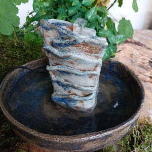 May include: A small, handcrafted ceramic fountain with a layered, textured design in shades of blue, brown, and white. The fountain sits in a round, shallow bowl filled with water. Green plants and moss surround the fountain, creating a natural aesthetic.