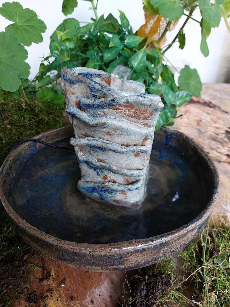 May include: A small, handcrafted ceramic fountain with a layered, textured design in shades of blue, brown, and white. The fountain sits in a round, shallow bowl filled with water. Green plants and moss surround the fountain, creating a natural aesthetic.