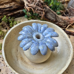 Könnte beinhalten: Ein Keramik-Räuchergefäß bestehend aus einer cremefarbenen Schale mit geprägtem Rand und einem zentralen, abnehmbaren blumenförmigen Halter. Die Blume hat glänzende, lavendelblaue Blütenblätter mit feinen gelben Sprenkeln und einer Öffnung.