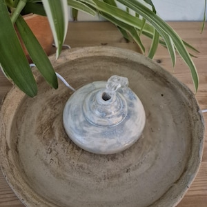 Può includere: Una fontana per animali in ceramica beige, dotata di un'ampia vasca strutturata e di un beccuccio centrale a cupola. La fontana presenta una smaltatura blu e bianca screziata sulla cupola centrale, con un cavo di alimentazione bianco laterale.