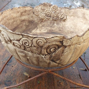 Puede incluir: Un macetero de cerámica rústico hecho a mano con un acabado texturizado de color blanco roto. El macetero presenta un diseño decorativo de patrones en espiral alrededor del borde. Se muestra en un simple soporte de metal.