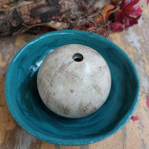May include: A beige, spherical ceramic object with a hole on top, in a teal ceramic bowl. The bowl is on a wooden surface. Decorative foliage and a piece of wood are in the background.