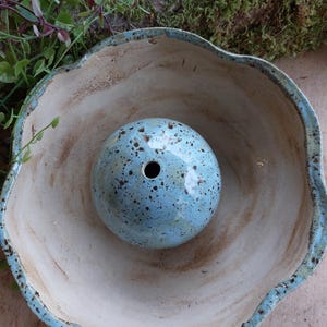 Könnte beinhalten: Handgefertigte Keramikschale mit hellblauem Rand und einer gesprenkelten blauen Kugel mit Loch. Die Schale hat einen welligen Rand und einen hellbeigen Innenraum. Die Kugel ist eine Vase oder ein dekoratives Objekt.