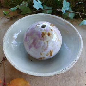 Könnte beinhalten: Eine kugelförmige Keramikvase mit einem einzelnen Loch, die in einer flachen, hellfarbigen Schale ruht. Die Kugel ist weiß mit Lavendel- und braunen Akzenten. Schale und Vase stehen auf einer Holzoberfläche, mit Grün im Hintergrund.
