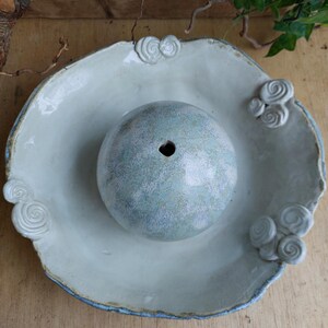May include: A pale blue ceramic bowl with a central sphere and decorative swirl accents. The bowl has a wide, irregular rim with a blue edge. The sphere has a hole at the top. The bowl is likely for decorative or functional use.