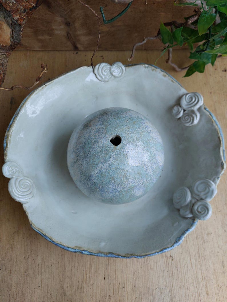 May include: A pale blue ceramic bowl with a central sphere and decorative swirl accents. The bowl has a wide, irregular rim with a blue edge. The sphere has a hole at the top. The bowl is likely for decorative or functional use.