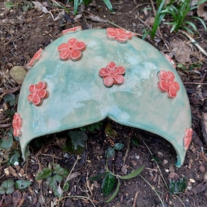 May include: A ceramic mushroom-shaped garden decoration with a teal glaze and red flower accents. The mushroom is hollow and has a small opening at the bottom.