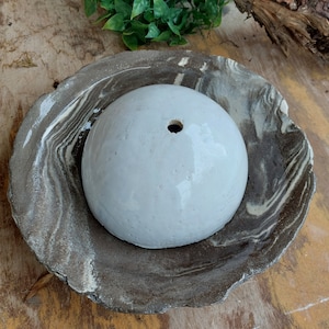 May include: A white, spherical ceramic planter with a single drainage hole, set within a marbled, brown and white ceramic dish. The dish has an organic, irregular shape. The items are on a wooden surface.