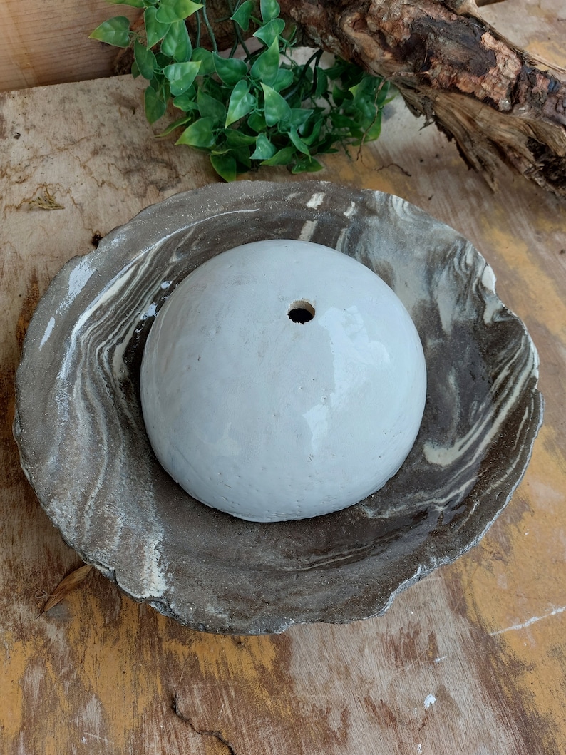 May include: A white, spherical ceramic planter with a single drainage hole, set within a marbled, brown and white ceramic dish. The dish has an organic, irregular shape. The items are on a wooden surface.