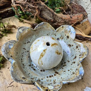 Könnte beinhalten: Eine handgefertigte Keramikschale in Blumenform mit einem passenden kugelförmigen Objekt. Die Schale hat eine hellblaue und braun gesprenkelte Glasur mit einem gewellten Rand. Die Kugel ist weiß mit braunen Streifen und einem Loch oben. Dekorative Keramik.