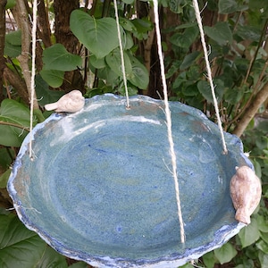 Può includere: Un bagno per uccelli in ceramica blu con un bordo ondulato appeso a due corde bianche. Due piccoli uccelli in ceramica sono appollaiati sul bordo del bagno.