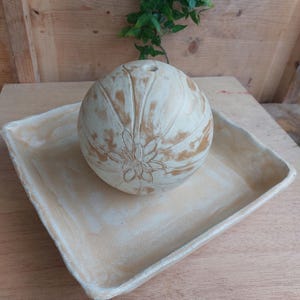 May include: A cream-coloured, spherical decorative object with a floral design, resting on a matching square tray. The sphere has a small opening at the top. The tray and sphere have a distressed, aged appearance.