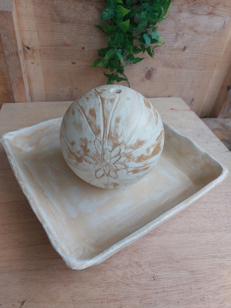 May include: A cream-coloured, spherical decorative object with a floral design, resting on a matching square tray. The sphere has a small opening at the top. The tray and sphere have a distressed, aged appearance.