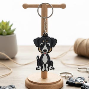 May include: A black and white dog keychain with a silver keyring is displayed on a wooden stand. Several similar keychains are scattered on a wooden surface. A small succulent plant in a gray pot and a spool of twine are also visible. A card reads "Handmade with Love."