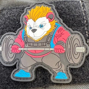 Könnte beinhalten: Ein Cartoon-Patch mit einem Löwen, der Gewichte hebt. Der Löwe trägt ein rot-blaues Gewichtheber-Outfit und hat ein weißes Gesicht mit blauen Augen. Der Patch ist aus besticktem Stoff und hat einen schwarzen Umriss.