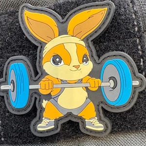 Può includere: Un coniglio dei cartoni animati che indossa un completo da sollevamento pesi e solleva un bilanciere con pesi blu e grigi. Il coniglio è arancione, bianco e marrone.