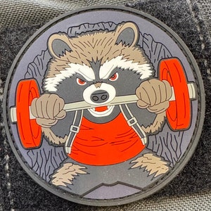 Könnte beinhalten: Ein gestickter Patch in Schwarz und Grau, der einen Waschbären zeigt, der ein rotes Tanktop trägt und eine Langhantel hebt. Der Waschbär hat einen grimmigen Ausdruck und hebt die Langhantel mit beiden Händen.