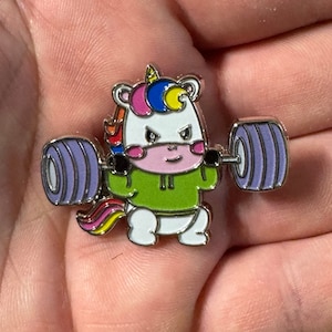 Könnte beinhalten: Ein weißes Einhorn mit einer Regenbogenmähne und einem Regenbogen-Schwanz hebt eine Langhantel mit violetten Gewichten. Das Einhorn trägt ein grünes Kapuzenoberteil und hat eine rosa Nase und rosa Wangen.