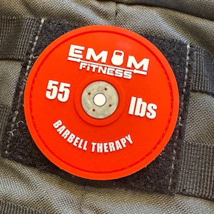 Puede incluir: Un parche redondo rojo con el texto "EM8M FITNESS" y "55 lbs BARBELL THERAPY" en blanco. El parche está adherido a una tela gris texturizada.