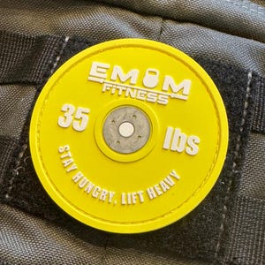 May include: A yellow weight plate patch with the text "EMOM FITNESS" and "35 lbs". The patch also includes the phrase "STAY HUNGRY, LIFT HEAVY". The patch is circular and has a silver center.